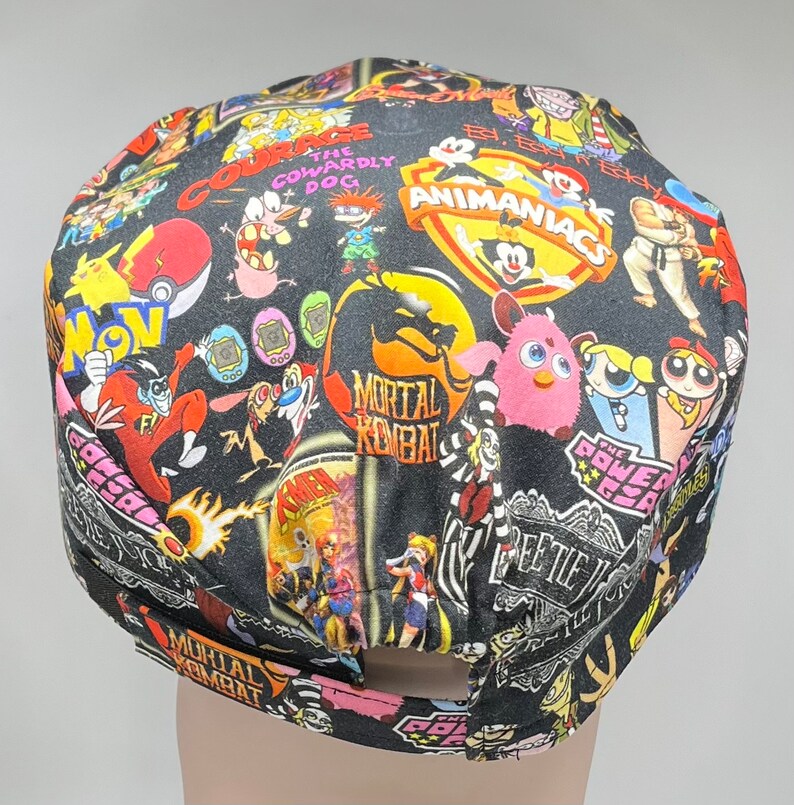 May include: A black scrub hat with a colorful print featuring cartoon characters from popular 90s shows, including 'The Powerpuff Girls', 'Animaniacs', 'Mortal Kombat', 'Courage the Cowardly Dog', and 'Dexter's Laboratory'.