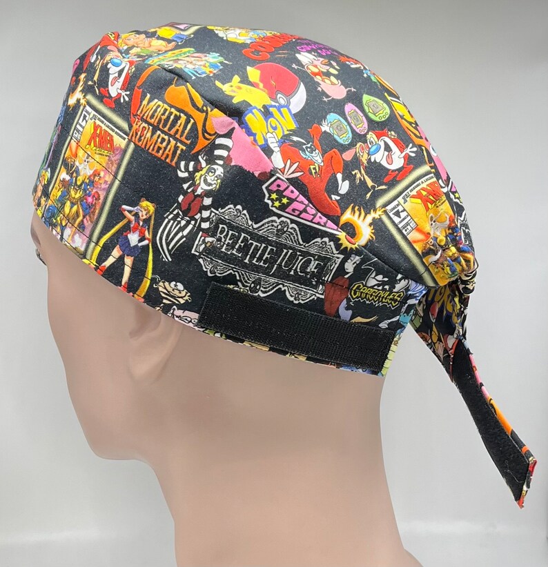 May include: A black scrub hat with a colorful print featuring cartoon characters, anime characters, and pop culture references. The hat has an adjustable strap and a long tail.