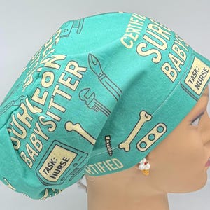 Könnte beinhalten: Eine türkisfarbene OP-Haube mit einem sich wiederholenden Muster aus medizinischen Werkzeugen und den Worten "SURGEON BABYSITTER" und "CERTIFIED NURSE". Die Haube ist für medizinisches Fachpersonal konzipiert und bietet eine bequeme Passform.