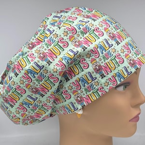 May include: A scrub cap with a light mint green background featuring a repeating pattern of nutcracker figures, small gold stars, and the text Y'ALL ARE NUTS in pink, purple, and blue lettering. The cap has a rounded, gathered crown design.