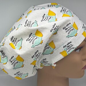 Peut inclure: Un bonnet de chirurgien blanc avec un motif répété de pélicans de dessin animé en bleu sarcelle, jaune et noir. Le texte "Stop acting like a disgruntled pelican" est imprimé autour des illustrations de pélicans. Le bonnet est pour les professionnels de la santé.
