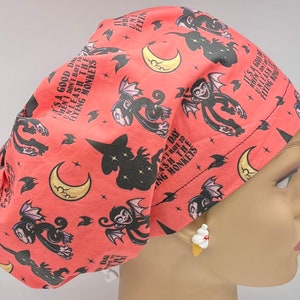 May include: Coral-colored surgical scrub cap with a repeating pattern of cartoon bats, moons, and witch silhouettes. Text reads "It's a good day when I don't have to unleash the flying monkeys." A fun and whimsical design.