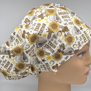 May include: A white scrub cap featuring a repeating pattern of yellow sunflowers, brown cowboy hats, horseshoes, and steam trains. The text "Take 'em to the TRAIN STATION" is printed in brown throughout the fabric, interspersed with small yellow stars.