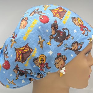 May include: A light blue surgical scrub hat with a whimsical pattern of monkeys, bananas, and a circus tent. The text "Monkey Business" is printed on the hat.