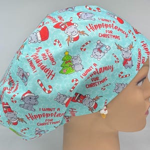 Könnte beinhalten: Eine hellblaue OP-Haube mit einem sich wiederholenden Muster aus Flusspferden in Weihnachtskleidung, Zuckerstangen und dem Text "I want a Hippopotamus for Christmas".