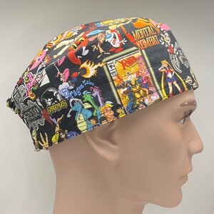 May include: A black scrub hat with a colorful print featuring cartoon characters and comic book covers. The print includes characters from 'Gargoyles', 'Dragon Tales', 'Mortal Kombat', 'Sailor Moon', and 'X-Men'.