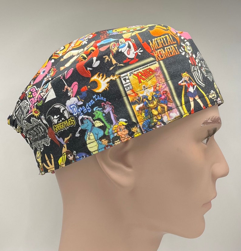 May include: A black scrub hat with a colorful print featuring cartoon characters and comic book covers. The print includes characters from 'Gargoyles', 'Dragon Tales', 'Mortal Kombat', 'Sailor Moon', and 'X-Men'.