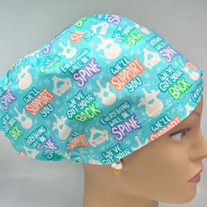 May include: A light blue scrub hat with a white spine graphic and the text "We've got your back" repeated in various colors. The hat is decorated with stars and the text "Everything will be spine" in a rainbow of colors.