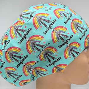 May include: A turquoise surgical scrub cap with a repeating pattern of skeletons, rainbows, and the text "SassyPants". The skeletons are wearing patterned pants and are positioned in front of colorful rainbows. The cap is designed for medical professionals.