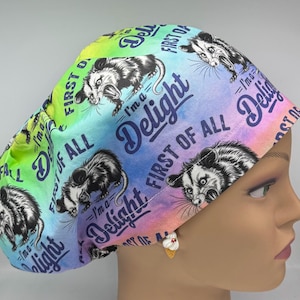 May include: A colorful scrub cap with a rainbow gradient background. The cap features opossum illustrations and the text "I'm a Delight, First of All". The cap is designed for medical professionals.