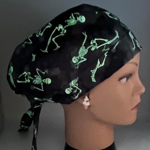 May include: Black scrub hat with a green glow-in-the-dark skeleton print. The hat has a tie at the back.