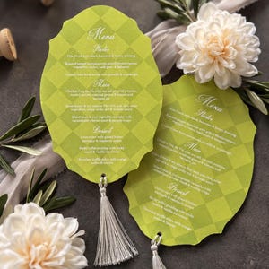 Puede incluir: Dos tarjetas de menú festoneadas a cuadros verde lima con texto en blanco, cada una adornada con una borla plateada en la parte inferior. Los menús enumeran secciones para Starter, Main y Dessert, rodeados de flores blancas y ramas de olivo.