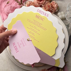 Può includere: Un menu a ventaglio con bordi smerlati composto da strati di cartoncino bianco, giallo e rosa chiaro. Il testo riporta DRINKS MENU, seguito da un elenco di portate e dai nomi DREW, AMELIE e MANUEL con la data 08 AUGUST 2026.