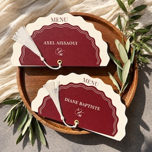 May include: Two semi-circular menu cards with scalloped edges, featuring a deep burgundy center and cream-colored border. Each card displays the word MENU, a name, and a monogram, accented by a white tassel attached through a gold-toned eyelet.