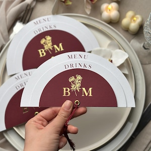 May include: A semi-circular menu and drinks card featuring a deep burgundy center with gold foil lettering that reads MENU DRINKS and the initials B M with a floral illustration. The card is held together by a gold eyelet and a red tassel.
