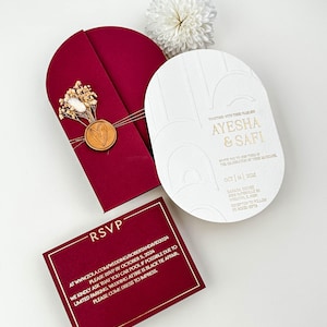 May include: Elegant wedding invitation suite featuring a burgundy arched invitation with a wax seal and dried flowers, a white oval invitation with gold text, and a burgundy RSVP card with gold trim. The invitations include the names Ayesha & Safi.