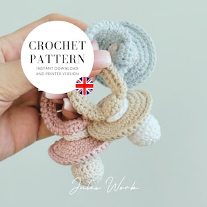May include: Three crocheted baby teethers in pink, beige, and light blue. The teethers are shaped like pacifiers with a ring handle. The teethers are held in a hand.