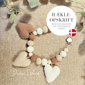 May include: A crochet pacifier clip with a heart motif. The clip features crochet balls in white, beige, and brown, interspersed with wooden beads. The clip has a metal clasp and a white heart-shaped crochet design. The text "HÆKLE-OPSKRIFT" is in a white circle.