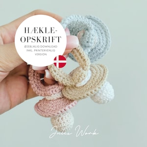 Puede incluir: Un conjunto de tres mordedores para bebés de crochet en rosa, beige y azul claro. Los mordedores tienen forma de chupete y tienen un anillo para un agarre fácil. La imagen incluye el texto "HÆKLE-OPSKRIFT ØJEBLIKLIG DOWNLOAD INKL. PRINTERVENLIG VERSION".