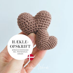 May include: Two crocheted brown hearts of different sizes. The larger heart is positioned above the smaller one. The image includes text in a white circle that reads "HÆKLEOPSKRIFT" and "ØJEBLIKLIG DOWNLOAD INKL. PRINTERVENLIG VERSION".