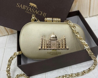 Op de afbeelding: Een goudkleurige, ovale clutch met een metalen architectonisch embleem van een gebouw met een koepel en minaretten. De tas is voorzien van een gouden kettinghengsel en draagt de tekst Sabyasachi Calcutta op de voorzijde en de doos.