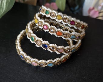 Könnte beinhalten: Ein Set aus fünf bunten Armreifen mit weißen Perlen und goldenen Akzenten. Jeder Armreif hat eine Reihe von ovalen Edelsteinen in verschiedenen Farben, darunter Blau, Grün, Gelb, Orange und Pink.