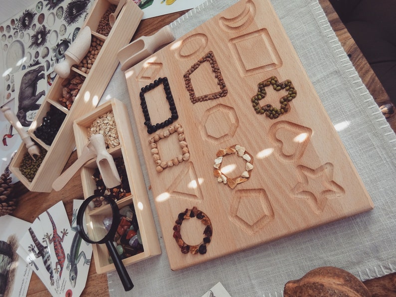 May include: A wooden board with various shapes cut out, filled with different types of beans and seeds. The board is made of light wood and has a natural finish. There are also several small wooden boxes filled with different types of beans and seeds.