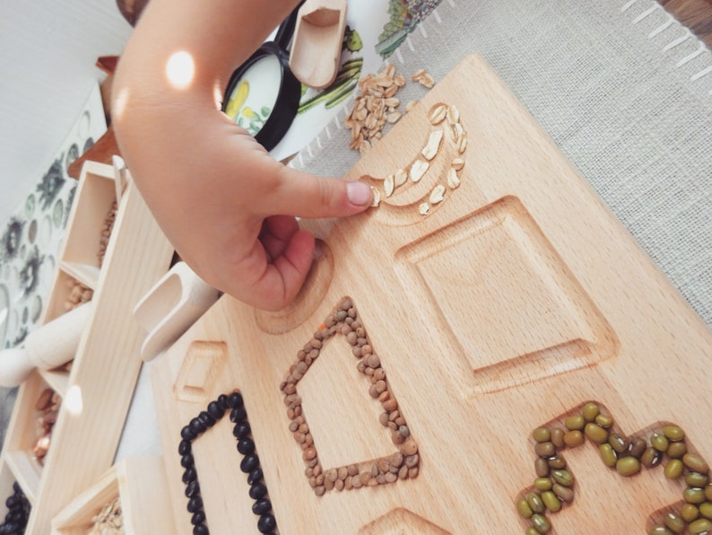 May include: A wooden tray with various shapes cut out, filled with different types of beans and grains. A child's hand is shown filling one of the shapes with oats.