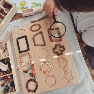May include: A wooden board with various shapes cut out, filled with different types of beans, seeds, and small stones. A child uses a magnifying glass to examine the details of the board.