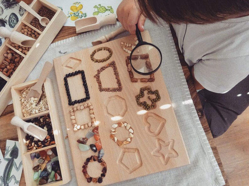 May include: A wooden board with various shapes cut out, filled with different types of beans, seeds, and small stones. A child uses a magnifying glass to examine the details of the board.