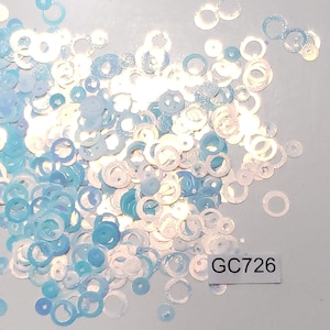 May include: A pile of iridescent blue and white glitter circles. The glitter is in the shape of a ring or donut. The glitter is scattered on a white surface. The text "GC726" is printed on a white label.