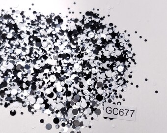 May include: A pile of black and silver glitter in various sizes. The glitter is scattered on a white surface. A white label with the text "GC677" is visible in the lower right corner of the image.