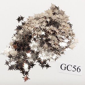 May include: A pile of silver glitter stars, perfect for crafting or decorating. The stars are scattered on a white background. The image also includes the text "GC56".