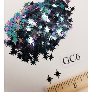 May include: A pile of iridescent black star-shaped glitter. The glitter is scattered on a white surface with a ruler and a white label with the text "GC6".
