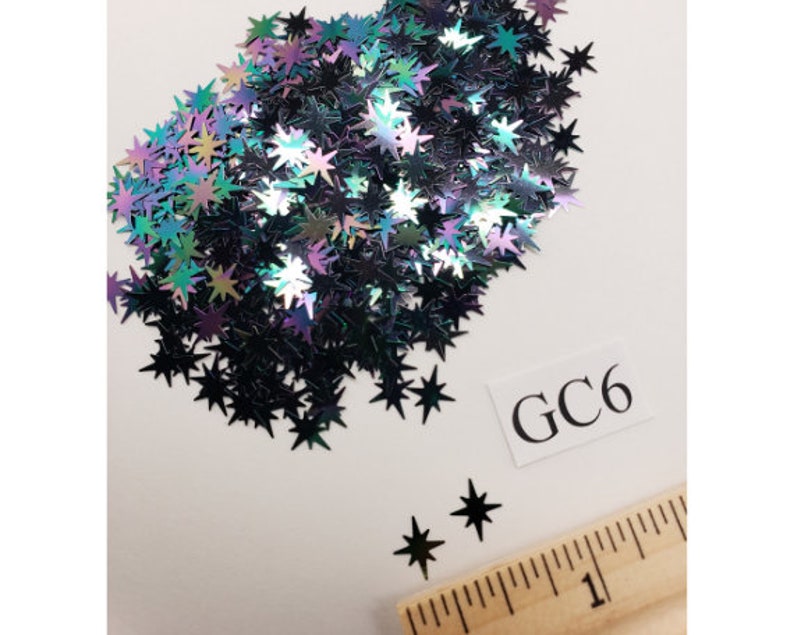 Puede incluir: Un mont&oacute;n de purpurina negra en forma de estrella iridiscente. La purpurina est&aacute; esparcida sobre una superficie blanca con una regla y una etiqueta blanca con el texto "GC6".