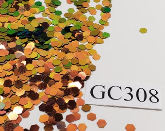 May include: A pile of iridescent green, gold, and orange glitter in the shape of hexagons. The glitter is scattered on a white surface. A white label with the text "GC308" is in the lower right corner of the image.