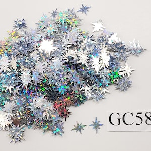May include: A pile of iridescent silver star-shaped glitter. The glitter is scattered on a white surface. The text "GC58" is written on a white label.