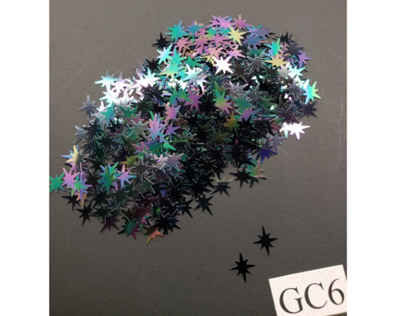 Puede incluir: Un mont&oacute;n de purpurina negra en forma de estrella iridiscente. La purpurina est&aacute; esparcida sobre una superficie gris. El texto "GC6" est&aacute; escrito en una etiqueta blanca en la esquina inferior derecha de la imagen.