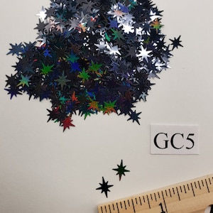 May include: A pile of iridescent black star-shaped glitter. The glitter is scattered on a white surface. The text "GC5" is written on a white label.