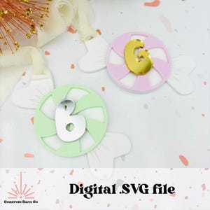 Può includere: Due ornamenti a forma di bastoncino di zucchero con un design a spirale. Uno è verde con un 6 argentato al centro e l'altro è rosa con una G dorata al centro. File SVG digitale.