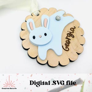 May include: A light blue bunny with pink ears is attached to a wooden circle with a scalloped edge. The circle is engraved with the name "Georgia".
