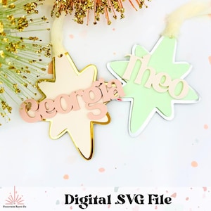 Puede incluir: Dos adornos en forma de estrella con los nombres "Georgia" y "Theo" en una fuente rosa suave. Uno es rosa claro con ribete dorado, el otro es verde claro con ribete plateado. El texto "Digital .SVG File" está en la parte inferior.