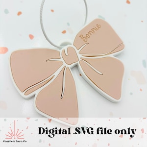 Puede incluir: Un lazo rosa con un contorno blanco y el nombre "Bonnie" grabado en él. El lazo está unido a un cordón plateado.