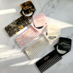 May include: A collection of four folding hair combs with attached circular mirrors. The combs feature tortoiseshell, pink marble, cream, and solid black finishes. Each piece is personalized with gold script names: Kate, Yasmin, Jolyn, Uray, Kristinan, and Raye.