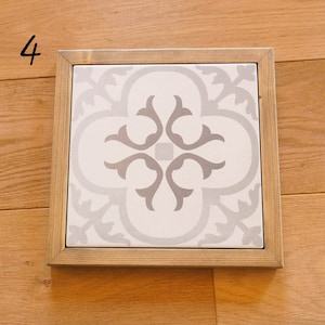 May include: A white ceramic tile with a gray floral pattern, framed in a light brown wood frame.