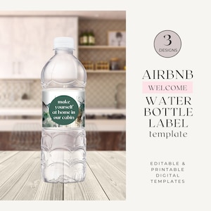 Puede incluir: Una botella de agua de plástico transparente con una etiqueta verde y blanca que dice "make yourself at home in our cabin". La etiqueta tiene un diseño de acuarela de una cabaña y un ciervo. La botella está sobre una superficie de madera.