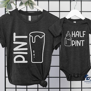 以下が含まれることがあります： 黒い格子に吊るされた、大人用とベビー用の2枚の濃いグレーのTシャツ。大人用Tシャツには、ビールグラスのグラフィックとともに「PINT」と書かれています。ベビー用ロンパースには、哺乳瓶のグラフィックとともに「HALF PINT」と書かれています。どちらのシャツも白です。