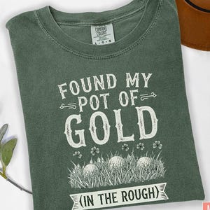 May include: Moss green t-shirt with the text "FOUND MY POT OF GOLD" in white, along with golf balls in grass. The shirt also has the text "(IN THE ROUGH)". The shirt is a Comfort Colors brand.