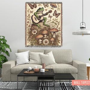 May include: A green frog playing a guitar with mushrooms and butterflies on a beige tapestry with a fringed border. The tapestry is hanging on a white wall above a beige couch.