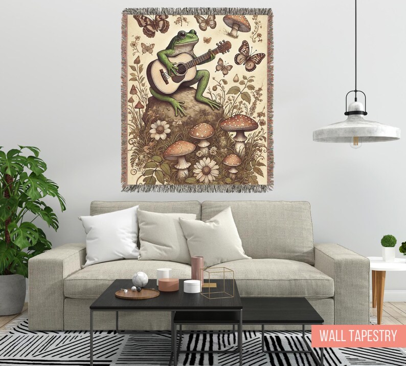 May include: A green frog playing a guitar with mushrooms and butterflies on a beige tapestry with a fringed border. The tapestry is hanging on a white wall above a beige couch.